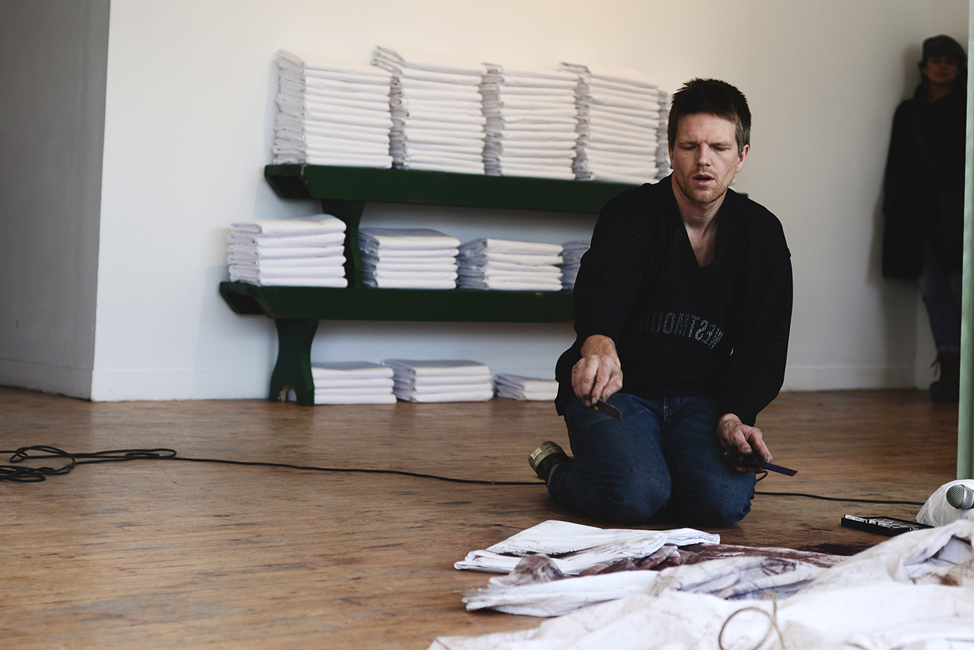 A man on his knees on the ground with stacks of folded white sheets on benches in the background.